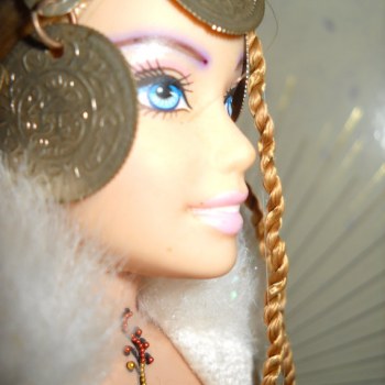 Azah wears a "Turkish Coat" of white with silver trim. Her dance attire is a band of ribbon top and a wrap skirt. She looks like a queen with her regal headress and fur neck collar. She also has some bindi adornment, and is naturally constructed of upcycled materials from dancers and barbies. 