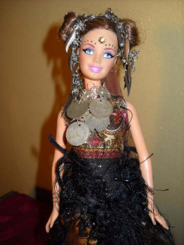 Tribal Diva Doll, Nalia is adorned and embellished with hair coiled and coifed, a jingly silver headdress and tribal coin necklace.  Her ethnic choli blouse is almost backless, she has a fringy beaded belly dance belt and flashy deep gold dance pants,  Her face bears tribal markings and she wears wrist gauntlets.